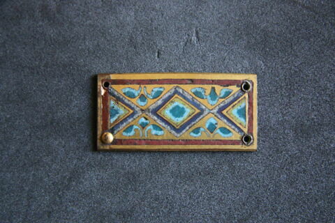 Plaquette rectangulaire : décorée d'un losange central émaillé et de deux demi-losanges bleu et vert sur fond réservé et doré ; bordure à filets d'émail rouge